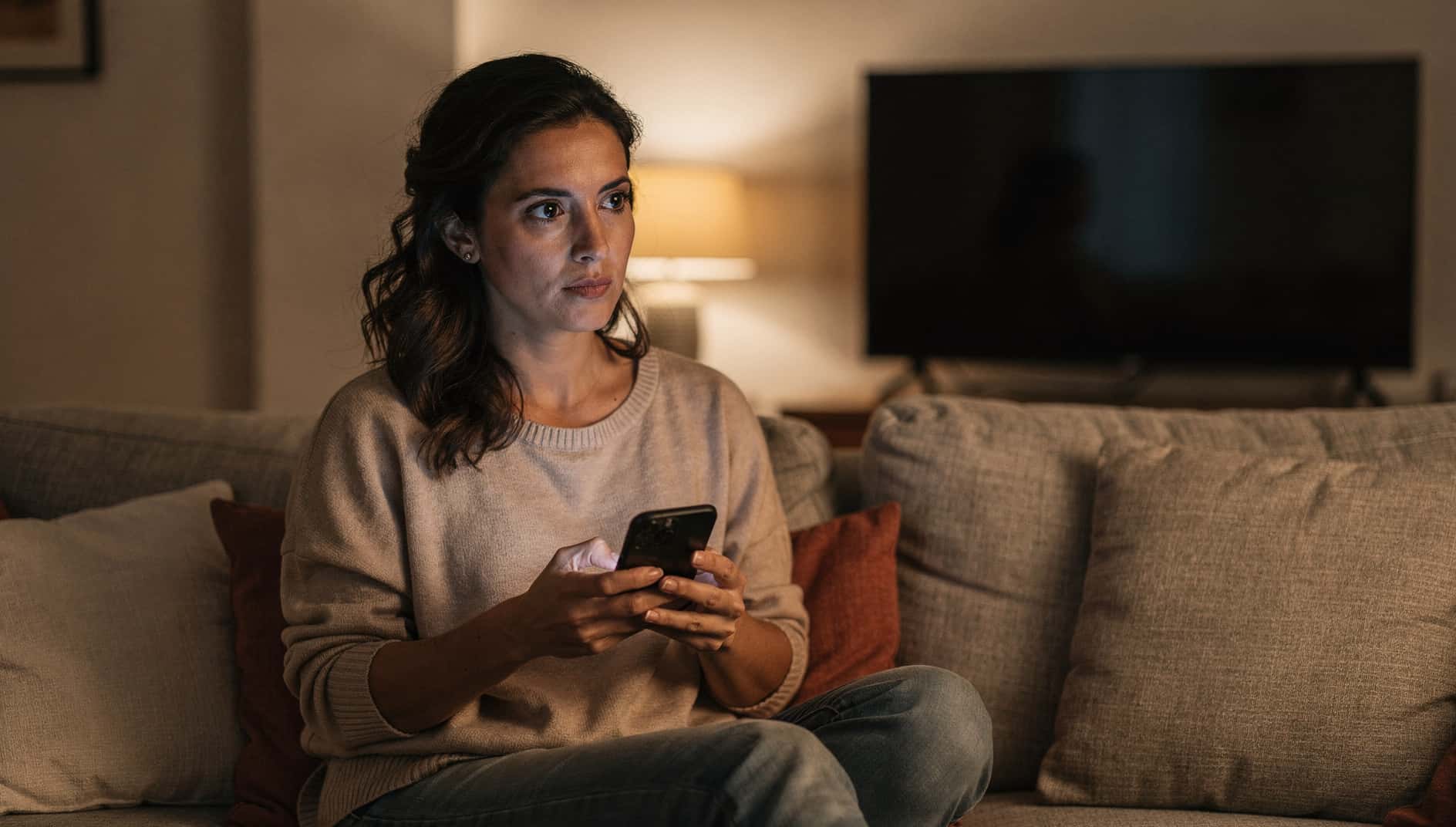 Mujer en el sofá con el móvil en la mano, pensando en cancelar una suscripción de streaming