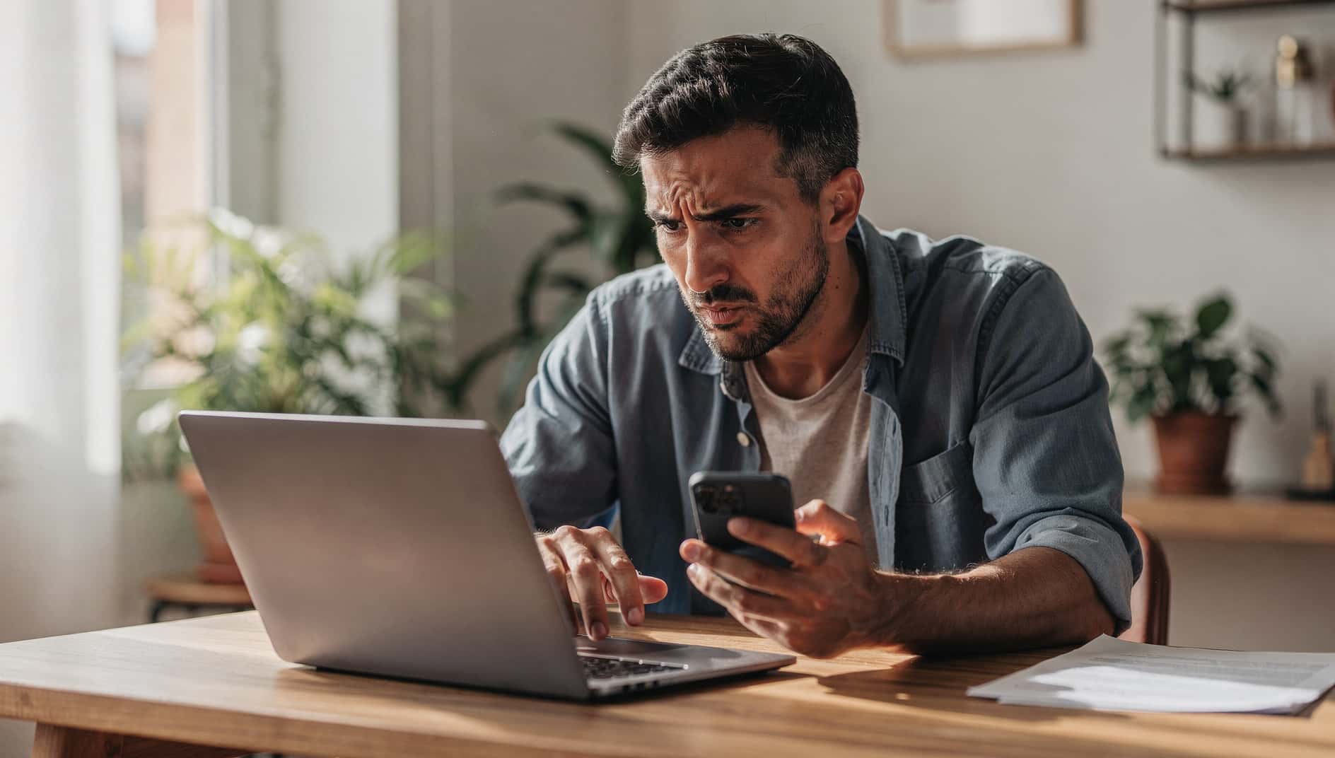 Hombre en su escritorio mirando el portátil y el móvil con gesto de frustración