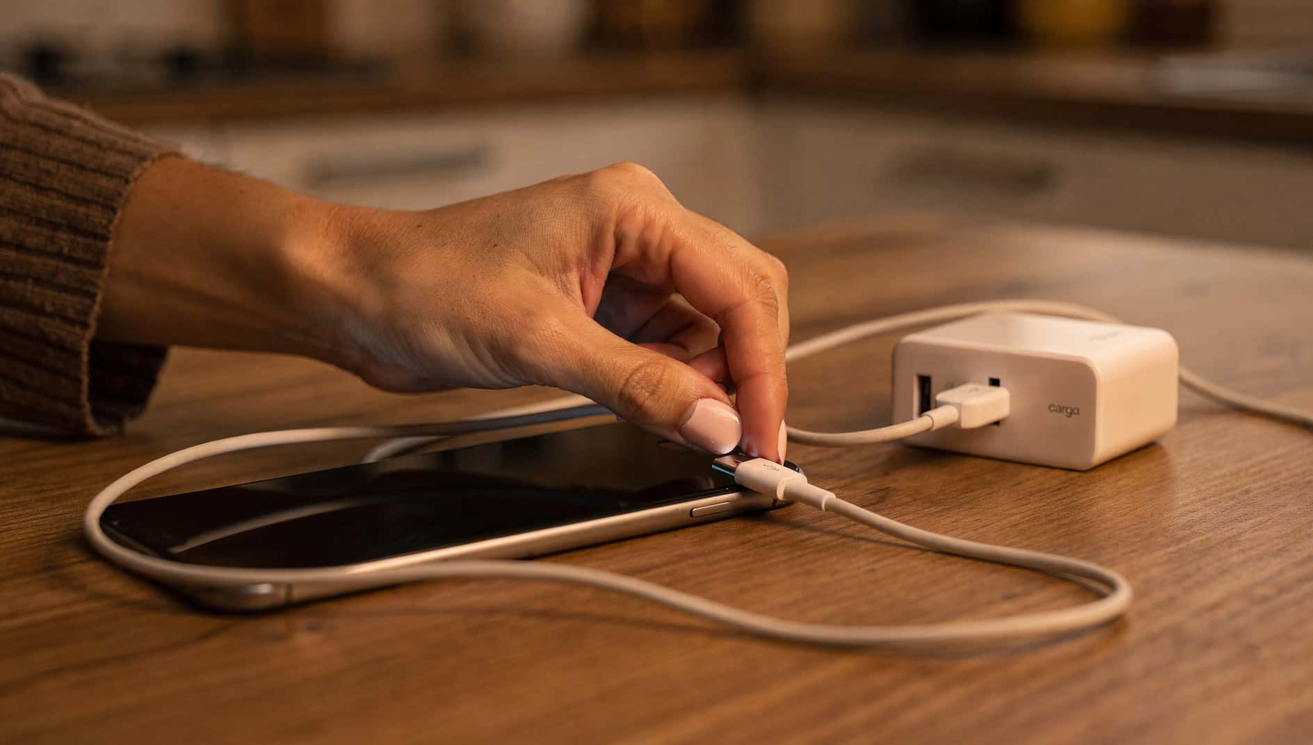 Mano conectando un cable de carga USB-C a un móvil sobre una mesa de cocina